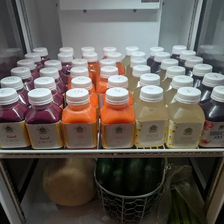 A cooler shelf stocked with fresh Cameron Juice bottles above a basket of melon and cucumbers