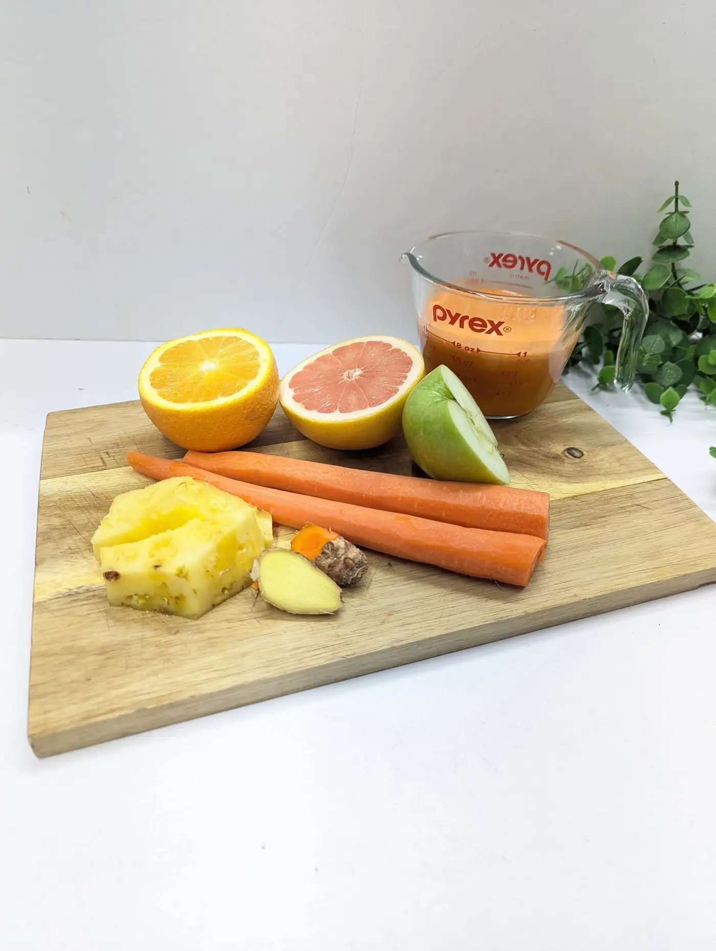 Ingredients for a citrus juice — orange, grapefruit, carrot, pineapple and apple — beside a cup of fresh juice