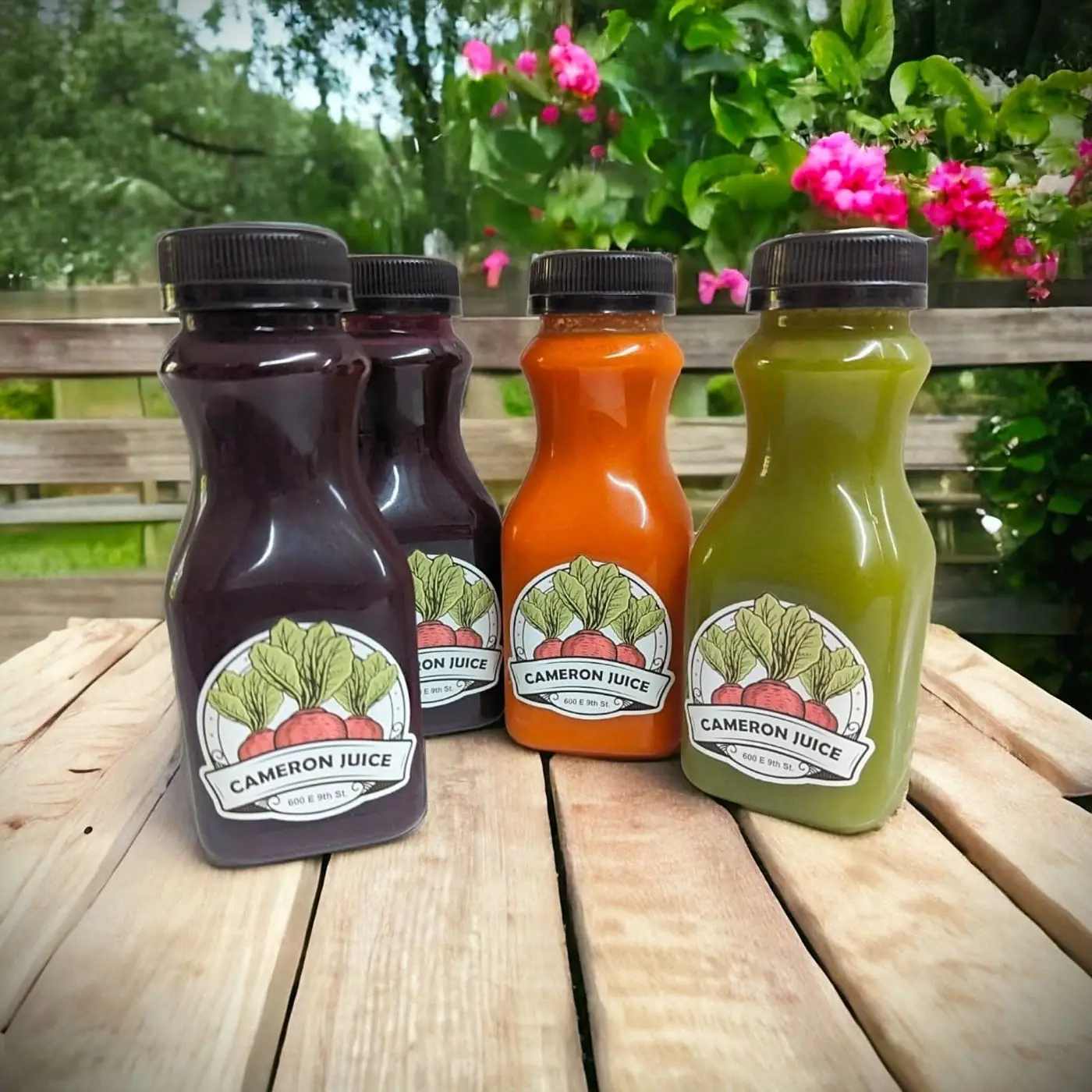 Four bottles of Cameron Juice cold-pressed juice on a wooden table outdoors with pink flowers behind