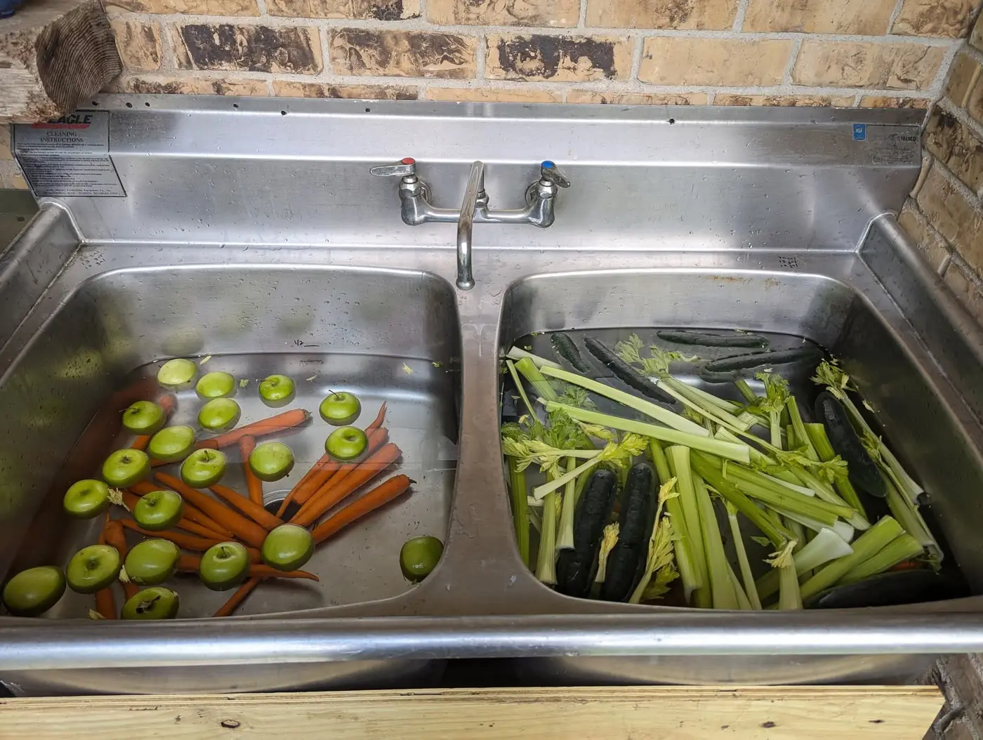Fresh carrots, green apples, celery and cucumbers being washed in a stainless prep sink